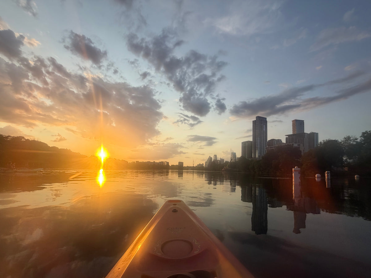 View from a kayak on Lady Bird Lake at sunset, with golden sunlight and the downtown Austin skyline reflected in calm water.